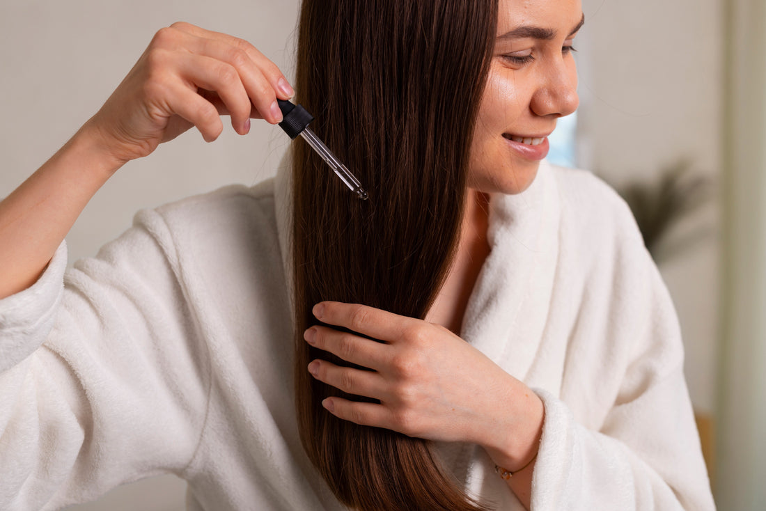 A candid, real-life photograph of a woman with brown hair performing an at-home hair repair routine. She is applying hair oil in the bathroom bathed in soft, natural daylight.