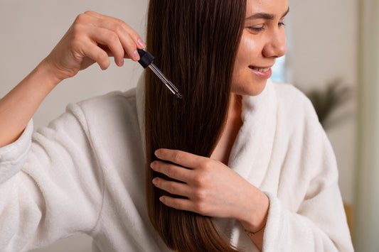A candid, real-life photograph of a woman with brown hair performing an at-home hair repair routine. She is applying hair oil in the bathroom bathed in soft, natural daylight.